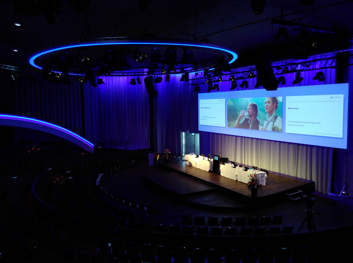 Grand auditorium avec scène éclairée sur laquelle se trouve un podium et au-dessus duquel est projeté un grand écran avec trois diapositives.